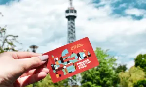 Dłoń trzyma czerwoną kartę "Prague Visitor Pass" na tle wieży widokowej i zielonych drzew.