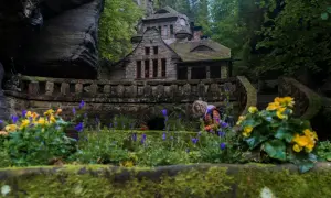 Kamienny dom w otoczeniu skał i zieleni w Parku Narodowym Czeska Szwajcaria. Kobieta pochyla się nad kwiatami.