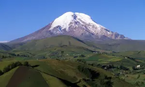 Pokryty śniegiem wulkan Chimborazo, jeden z najwyższych gór świata, góruje nad zielonymi, pofałdowanymi wzgórzami z polami uprawnymi i domami.
