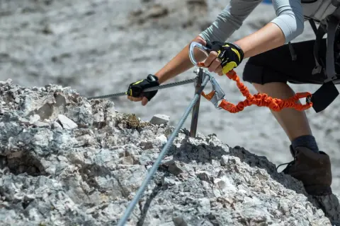 Ręce w rękawiczkach pewnie trzymają linę na via ferrata Słowacja. Pomarańczowa lonża z karabinkami zapewnia bezpieczeństwo.