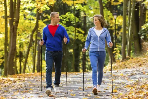 Para spaceruje po jesiennym parku, uprawiając nordic walking. Liście tworzą kolorowy dywan.