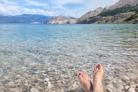 Stopy w klapkach na kamienistej plaży, krystalicznie czysta woda i górzyste wybrzeże. Raj na wyspie Krk.