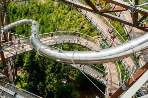 Drewniana ścieżka i metalowa zjeżdżalnia na Sky Walk Dolni Morava wśród zielonych drzew.
