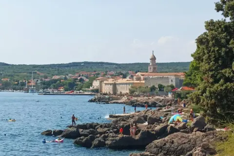 Słoneczna plaża na wyspie Krk, z kamienistym wybrzeżem, turkusowym morzem i miasteczkiem w tle. Wiele osób korzysta z uroków wyspy Krk, zwiedzając jej atrakcje.