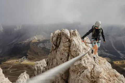 Kobieta na via ferrata polska pokonuje skalną grań, w tle górzysty krajobraz z krętymi drogami.