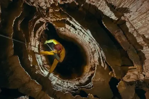 Speleolog w żółtym kombinezonie zjazduje w głąb skalnej otchłani, eksplorując potencjalnie najgłębszą jaskinię na świecie.