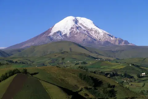 Pokryty śniegiem wulkan Chimborazo, jeden z najwyższych gór świata, góruje nad zielonymi, pofałdowanymi wzgórzami z polami uprawnymi i domami.
