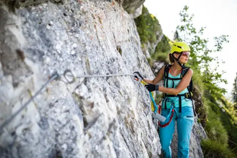 Kobieta w kasku i sprzęcie wspinaczkowym pokonuje via ferratę. To bezpieczna trasa w górach, gdzie ferrata co to jest, pomaga lina i stopnie.