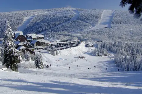 Ośnieżone stoki i lasy wokół ośrodka cervenohorske sedlo ski. Narciarze szusują po łagodnych zboczach, a w tle widać parking i zabudowania.
