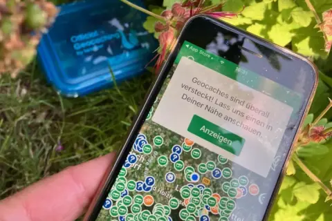 Smartfon pokazuje mapę z ukrytymi skarbami. To właśnie geocaching co to za zabawa! W tle niebieskie pudełko z napisem "Official Geocaching".