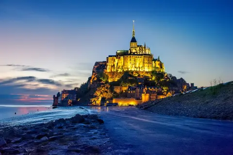 Mont Saint-Michel, jeden z najpiękniejszych zamków w Europie, lśni w blasku wieczornych świateł na tle błękitnego nieba.