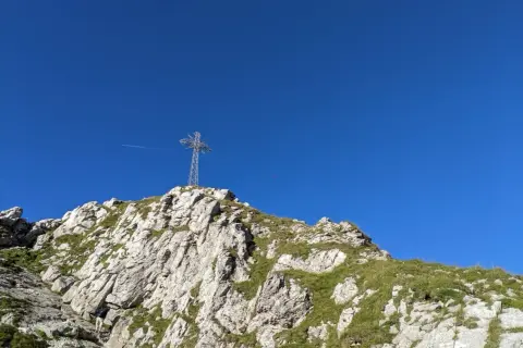 Giewont, śpiący rycerz, wznosi się ku błękitnemu niebu. Na szczycie stoi krzyż, a pod nim skaliste zbocza porośnięte trawą.