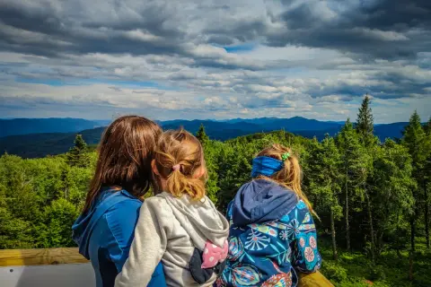 Rodzina podziwia widok z wieży widokowej. W dole rozciąga się zielona dolina, a w oddali piętrzą się góry. To idealne miejsce na rodzinną wycieczkę, gdzie można podziwiać piękne krajobrazy i cieszyć się wspólnym czasem. Trasa widokowa jest łatwo dostęp...