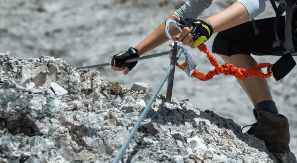 Ręce w rękawiczkach pewnie trzymają linę na via ferrata Słowacja. Pomarańczowa lonża z karabinkami zapewnia bezpieczeństwo.