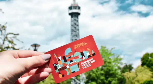 Dłoń trzyma czerwoną kartę "Prague Visitor Pass" na tle wieży widokowej i zielonych drzew.