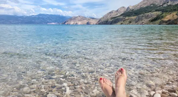 Stopy w klapkach na kamienistej plaży, krystalicznie czysta woda i górzyste wybrzeże. Raj na wyspie Krk.