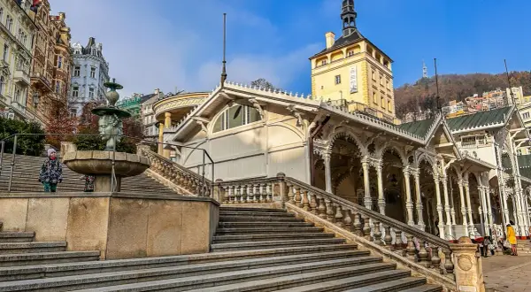 Schody prowadzące do Kolumnady w Karlowych Warach, z fontanną i zabytkowymi budynkami w tle.