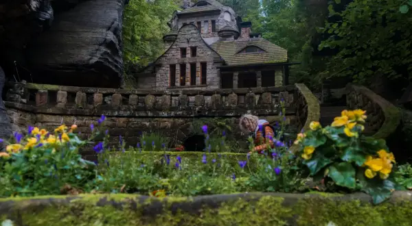 Kamienny dom w otoczeniu skał i zieleni w Parku Narodowym Czeska Szwajcaria. Kobieta pochyla się nad kwiatami.