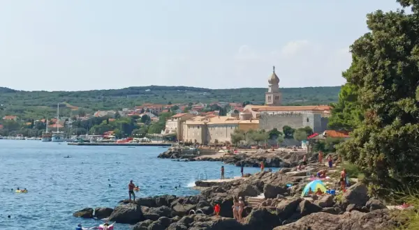 Słoneczna plaża na wyspie Krk, z kamienistym wybrzeżem, turkusowym morzem i miasteczkiem w tle. Wiele osób korzysta z uroków wyspy Krk, zwiedzając jej atrakcje.