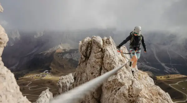 Kobieta na via ferrata polska pokonuje skalną grań, w tle górzysty krajobraz z krętymi drogami.