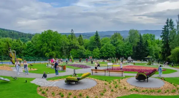 Kolorowy park z rzeźbami z kwiatów i ludźmi spacerującymi po alejkach. W oddali widać zalesione wzgórza. Ile się idzie, tyle piękna!