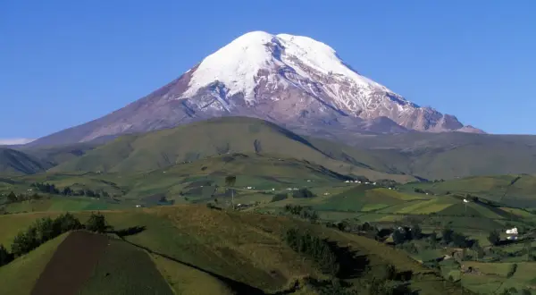 Pokryty śniegiem wulkan Chimborazo, jeden z najwyższych gór świata, góruje nad zielonymi, pofałdowanymi wzgórzami z polami uprawnymi i domami.