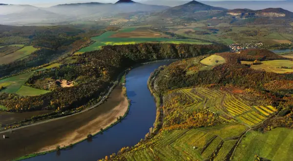 Malowniczy krajobraz z krętą rzeką, polami i lasami w jesiennych barwach. W oddali widać góry, charakterystyczne dla krajobrazów **rzeki w Czechach**.