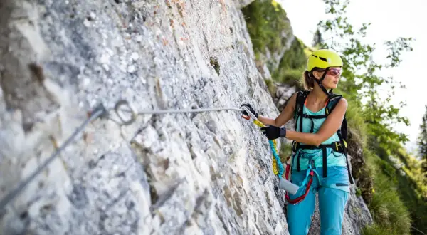 Kobieta w kasku i sprzęcie wspinaczkowym pokonuje via ferratę. To bezpieczna trasa w górach, gdzie ferrata co to jest, pomaga lina i stopnie.