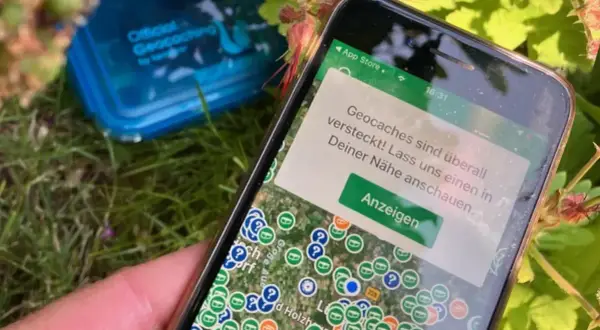 Smartfon pokazuje mapę z ukrytymi skarbami. To właśnie geocaching co to za zabawa! W tle niebieskie pudełko z napisem "Official Geocaching".