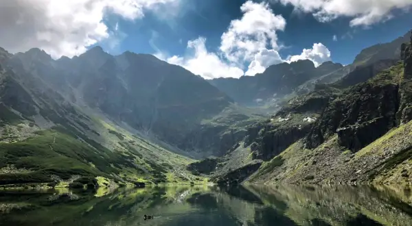 Malowniczy Czarny Staw Gąsienicowy, odbijający się w tafli wody, otoczony stromymi, skalistymi górami. Trasa widoczna na zboczu.