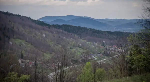Widok na dolinę z zabudowaniami, lasami i pasmem górskim w tle. Widać tu ćwilin z Jurkowa, wiosenną zieleń i chmury na niebie.