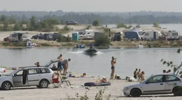 Ludzie odpoczywają nad jeziorem, niektórzy kąpią się, inni opalają na plaży. W tle widać kampery i samochody, a na wodzie pływa motorówka. To idealne miejsce na wakacje, jak na pogoria 4.