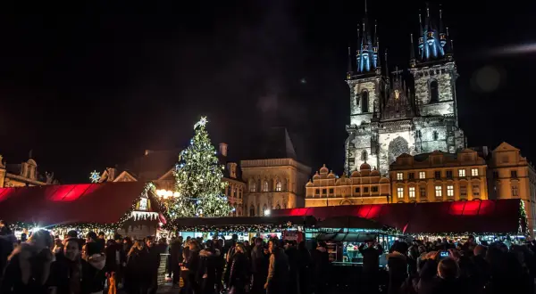 Tłum ludzi na jarmarku bożonarodzeniowym w Pradze, w tle iluminowana choinka i zabytkowe budynki.