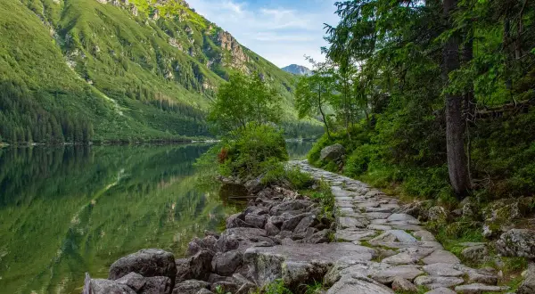 Kamienista ścieżka wzdłuż Morskiego Oka, z zielonymi zboczami gór i odbijającymi się w wodzie chmurami.