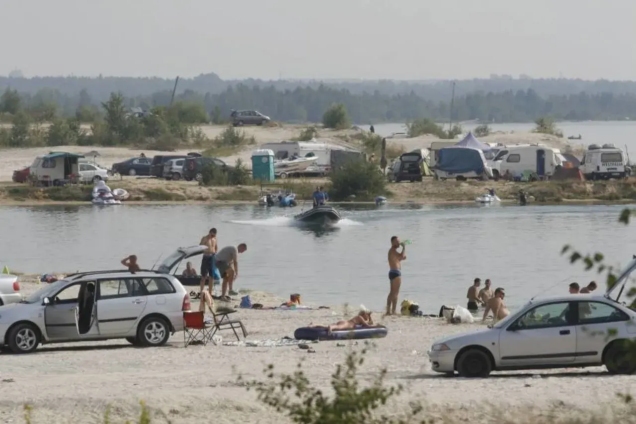 Ludzie odpoczywają nad jeziorem, niektórzy kąpią się, inni opalają na plaży. W tle widać kampery i samochody, a na wodzie pływa motorówka. To idealne miejsce na wakacje, jak na pogoria 4.