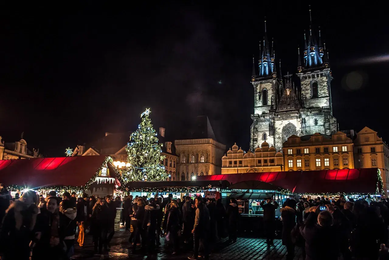 Tłum ludzi na jarmarku bożonarodzeniowym w Pradze, w tle iluminowana choinka i zabytkowe budynki.