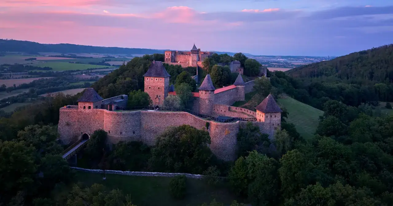 Jak wyglądają Czechy? Majestatyczny zamek na wzg&oacute;rzu, otoczony zielenią, w blasku r&oacute;żowego zmierzchu.