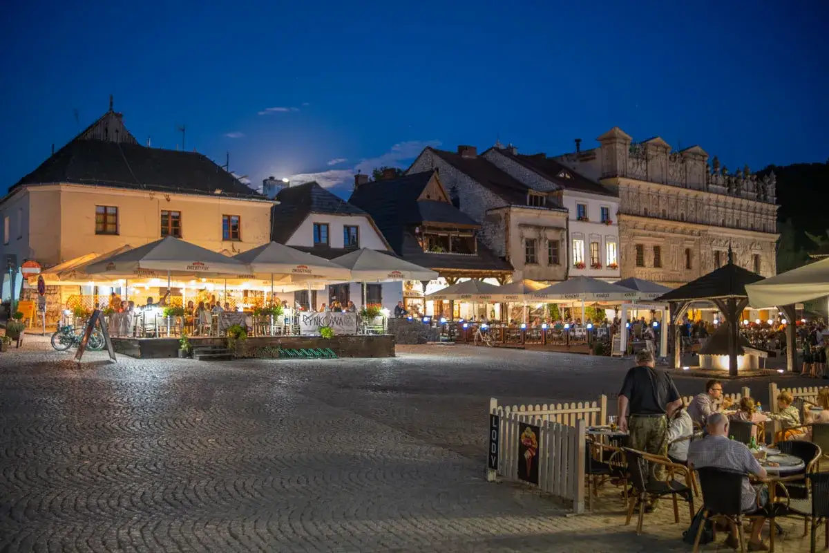 Wieczorny Rynek w Kazimierzu Dolnym tętni życiem. Stoliki restauracyjne pod parasolami, oświetlone kamienice i gwar rozm&oacute;w tworzą niepowtarzalny klimat. Kazimierz Dolny atrakcje wieczorem.