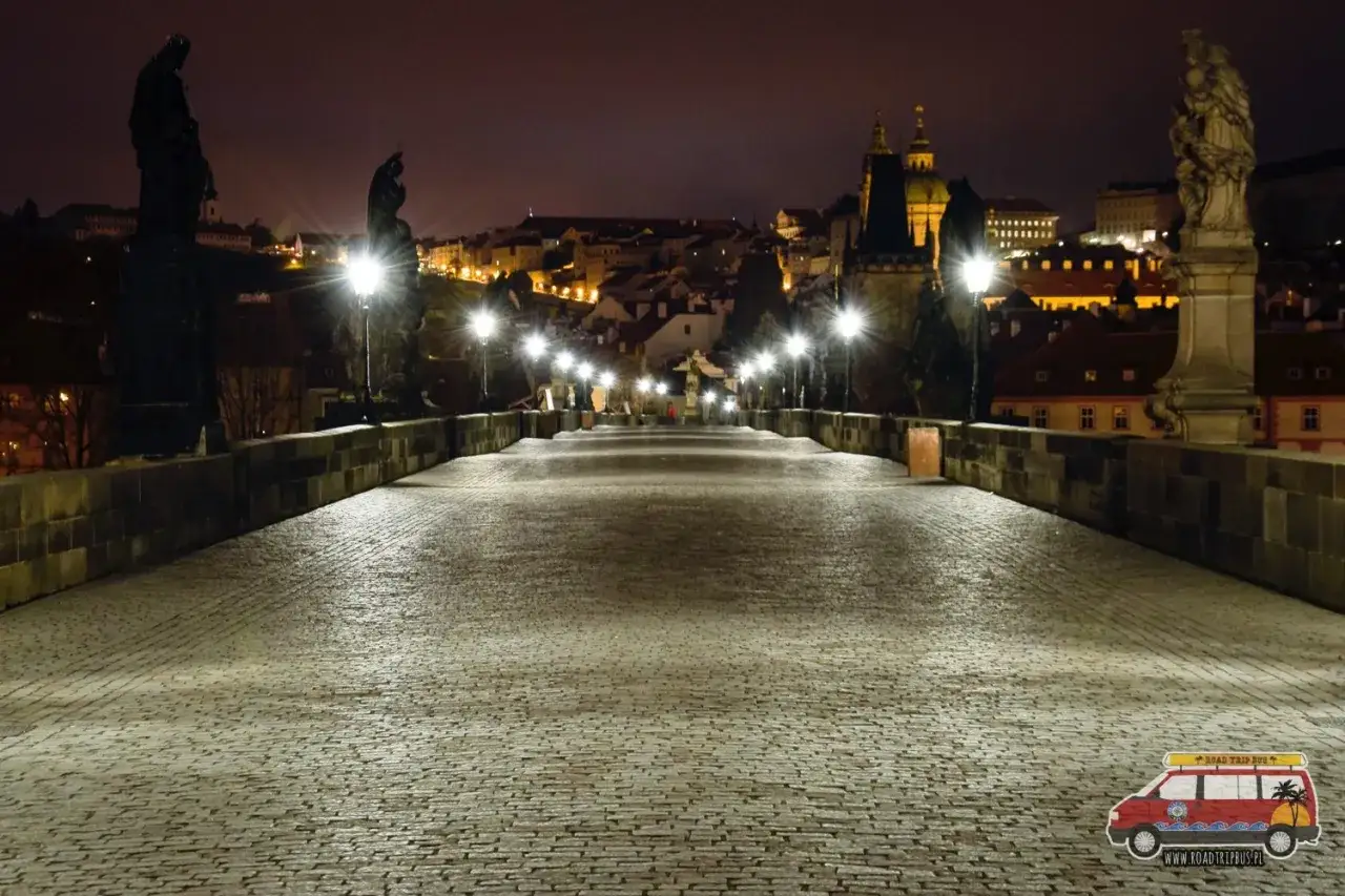 Kamienny most w nocy, oświetlony latarniami, z widokiem na praskie kamienice. Idealne miejsce, by poczuć atmosferę miasta, co warto zobaczyć w drodze do Pragi.