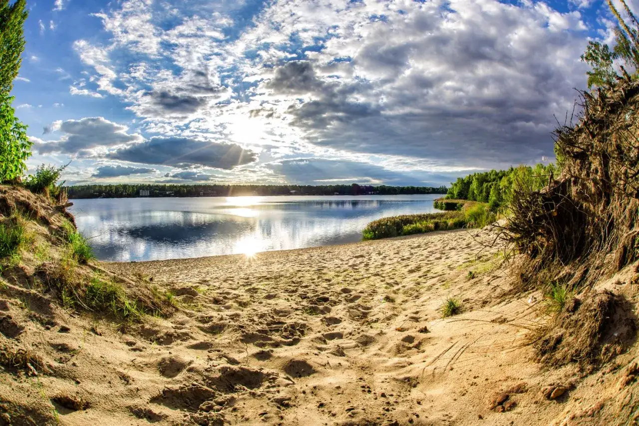 Piękny widok na jezioro z piaszczystą plażą i drzewami. Słońce przebija się przez chmury, tworząc refleksy na wodzie. Idealne miejsce na odpoczynek, jak na pogoria 4.