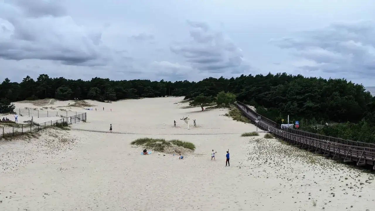 Spacer po piaszczystych wydmach w Dźwirzynie. Ludzie odpoczywają i grają w siatk&oacute;wkę plażową. Drewniana kładka prowadzi przez wydmy do lasu.