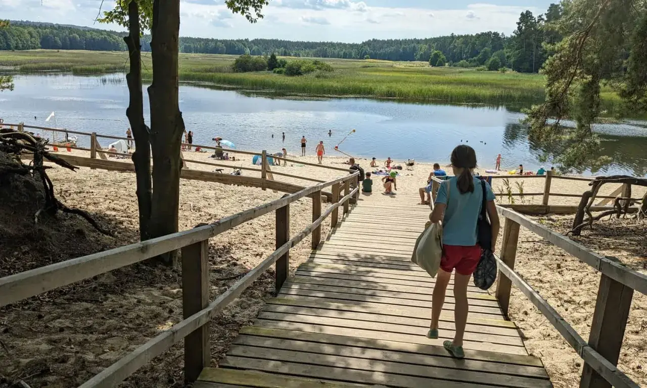 Dziewczynka schodzi drewnianą trasą na piaszczystą plażę nad jezioro. W oddali widać ludzi kąpiących się w wodzie i zieleń. Stawy Echo to idealne miejsce na letni wypoczynek.