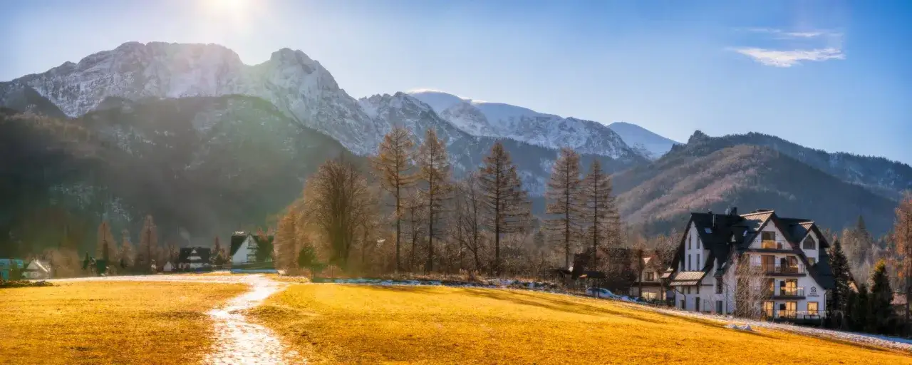 Zimowy krajobraz z górami w tle. Widać białe budynki, drzewa i złote pole. Gdzie jest napis Zakopane?