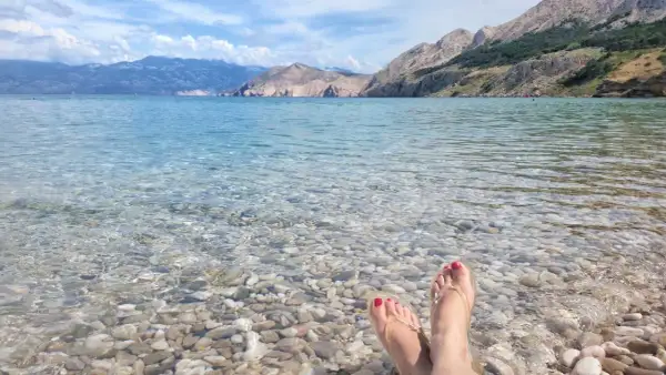 Stopy w klapkach na kamienistej plaży, krystalicznie czysta woda i górzyste wybrzeże. Raj na wyspie Krk.