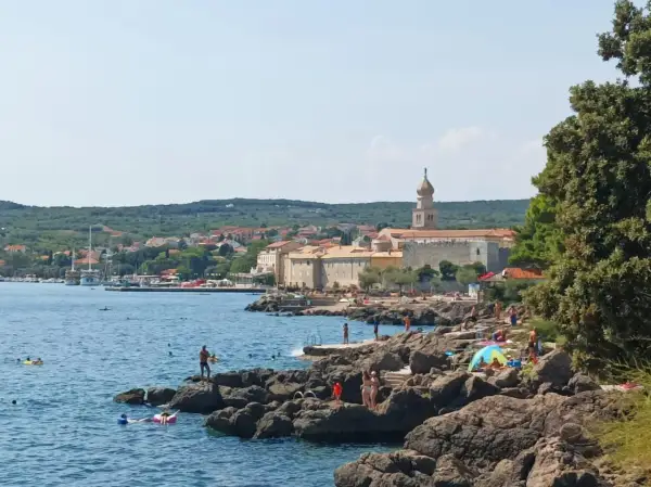 Słoneczna plaża na wyspie Krk, z kamienistym wybrzeżem, turkusowym morzem i miasteczkiem w tle. Wiele osób korzysta z uroków wyspy Krk, zwiedzając jej atrakcje.
