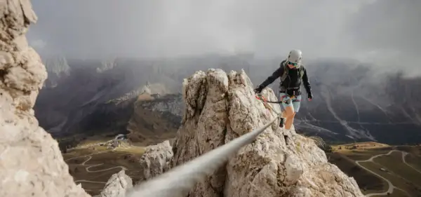 Kobieta na via ferrata polska pokonuje skalną grań, w tle górzysty krajobraz z krętymi drogami.