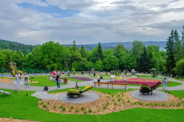 Kolorowy park z rzeźbami z kwiatów i ludźmi spacerującymi po alejkach. W oddali widać zalesione wzgórza. Ile się idzie, tyle piękna!