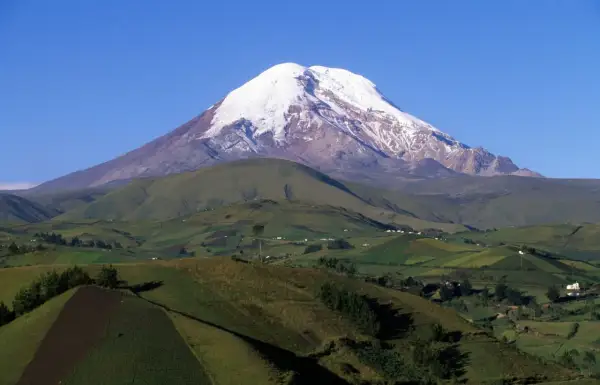 Pokryty śniegiem wulkan Chimborazo, jeden z najwyższych gór świata, góruje nad zielonymi, pofałdowanymi wzgórzami z polami uprawnymi i domami.