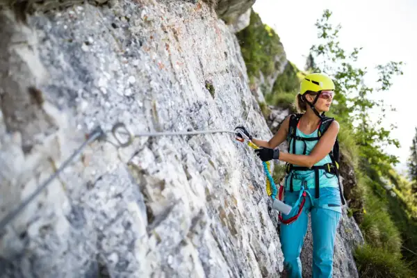 Kobieta w kasku i sprzęcie wspinaczkowym pokonuje via ferratę. To bezpieczna trasa w górach, gdzie ferrata co to jest, pomaga lina i stopnie.