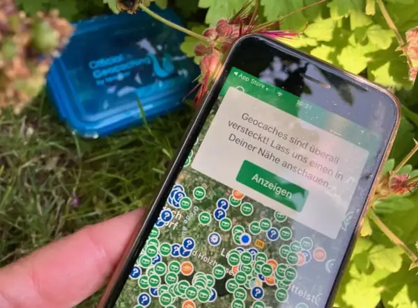 Smartfon pokazuje mapę z ukrytymi skarbami. To właśnie geocaching co to za zabawa! W tle niebieskie pudełko z napisem "Official Geocaching".